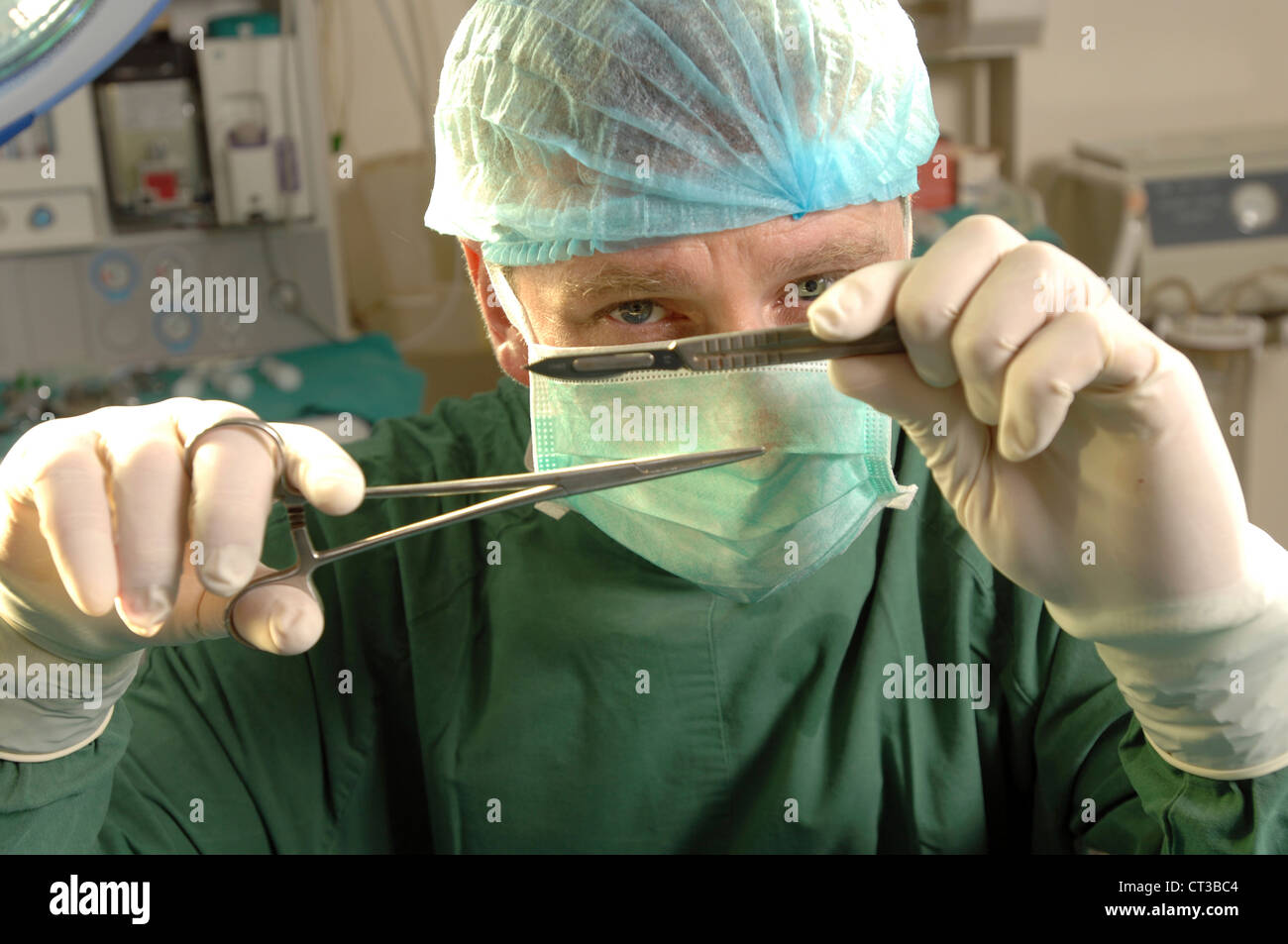 A masked surgeon holding up a pair of forceps and a scalpel in his gloved hands. Stock Photo