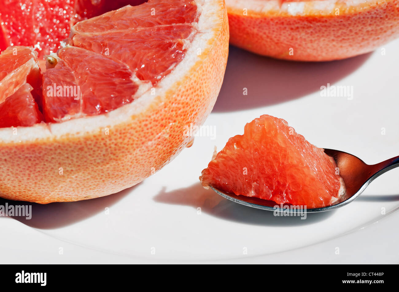 A bite of fresh juicy grapefruit on a spoon rests on a plate next to a halved grapefruit. Stock Photo