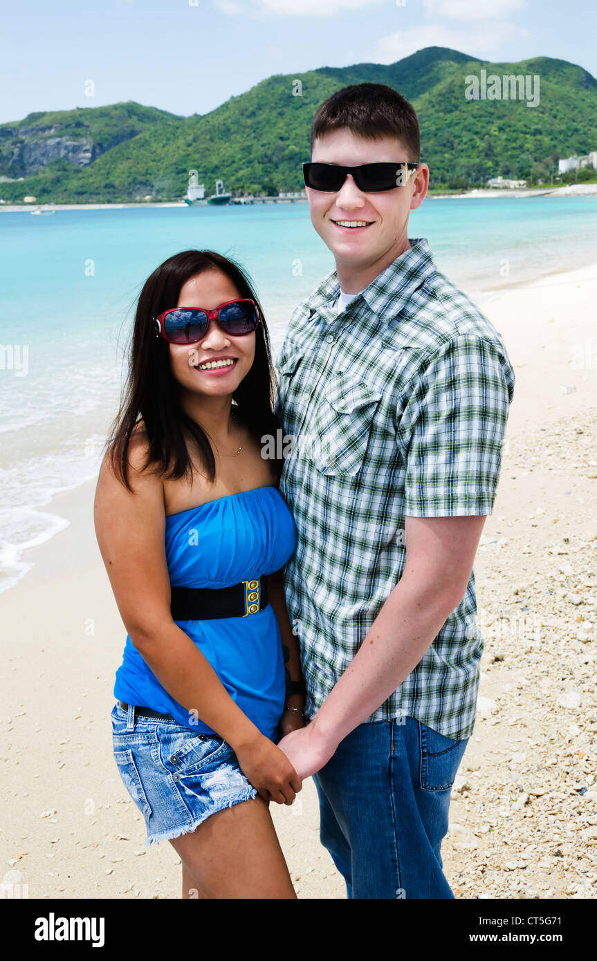 Young adult mixed race couple holds hands and smiles for the camera on a beautiful beach in Okinawa, Japan. Stock Photo
