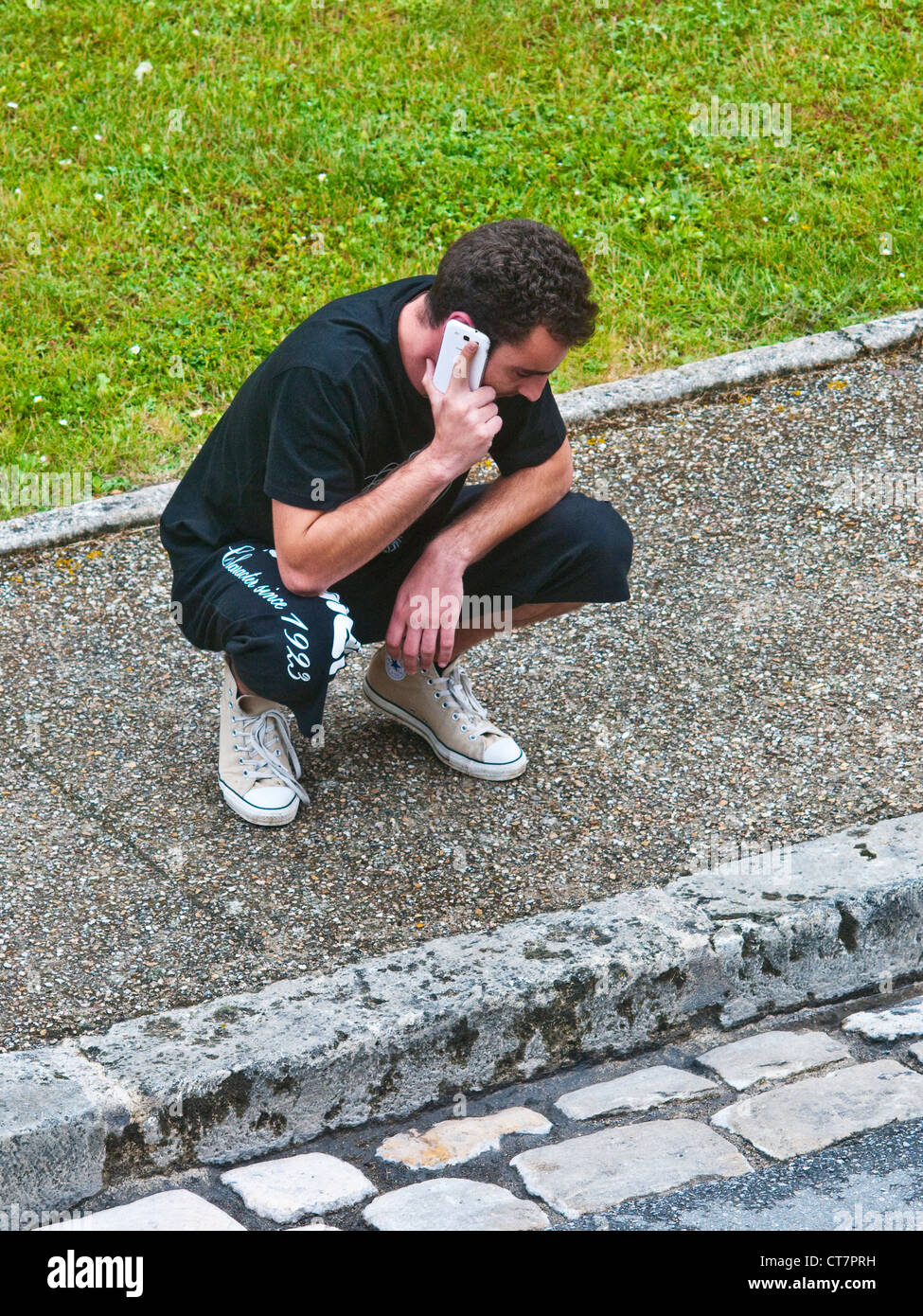 Younger man with telephone crouching on pavement - France. Stock Photo