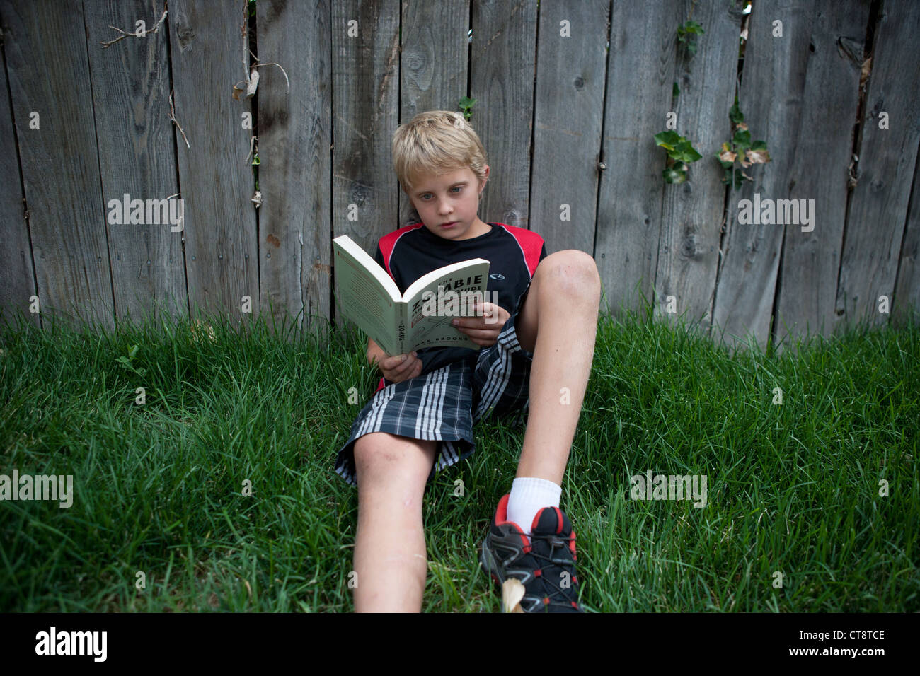 Ten year old boy reading a book, sitting in the grass and leaning against fence. Stock Photo