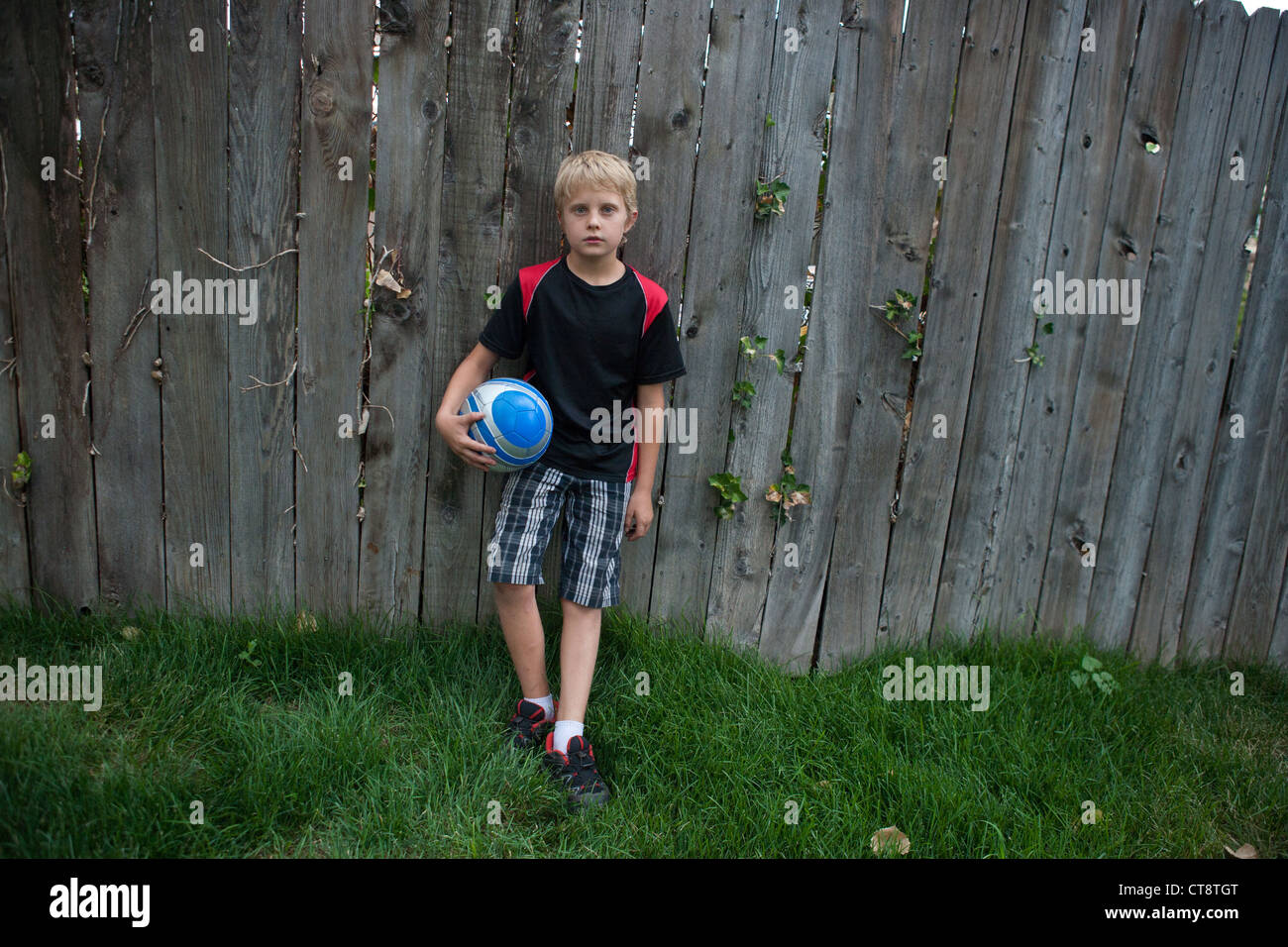 Ten year old boy holding a soccer ball and leaning against a wood fence