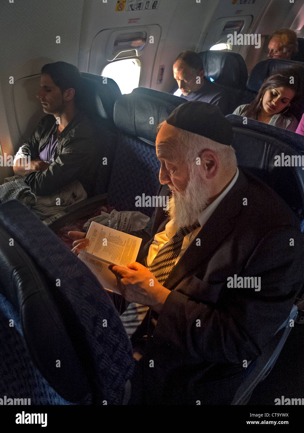 Wearing a yarmulke skull cap, an bearded Orthodox Jew reads from a book in Hebrew on an airliner while passengers sleep. Stock Photo