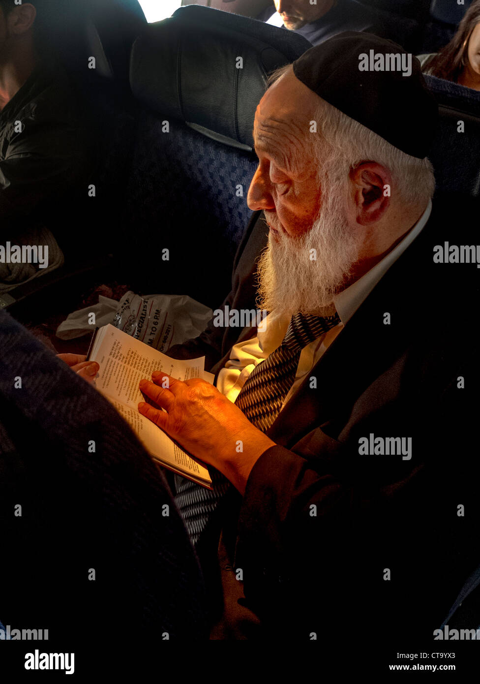 Wearing a yarmulke skull cap, an bearded Orthodox Jew reads from a book in Hebrew on an airliner while passengers sleep. Stock Photo