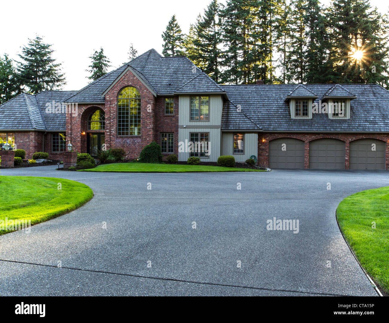 Driveway to large brick and cedar home with trees and sunlight in background Stock Photo