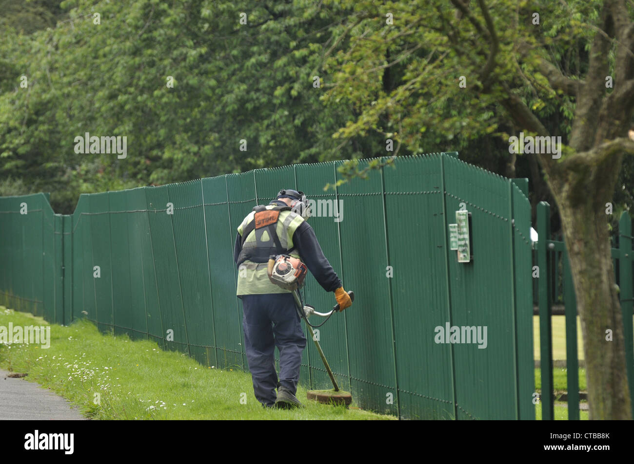 Council worker strimming grass in local park by fence Stock Photo