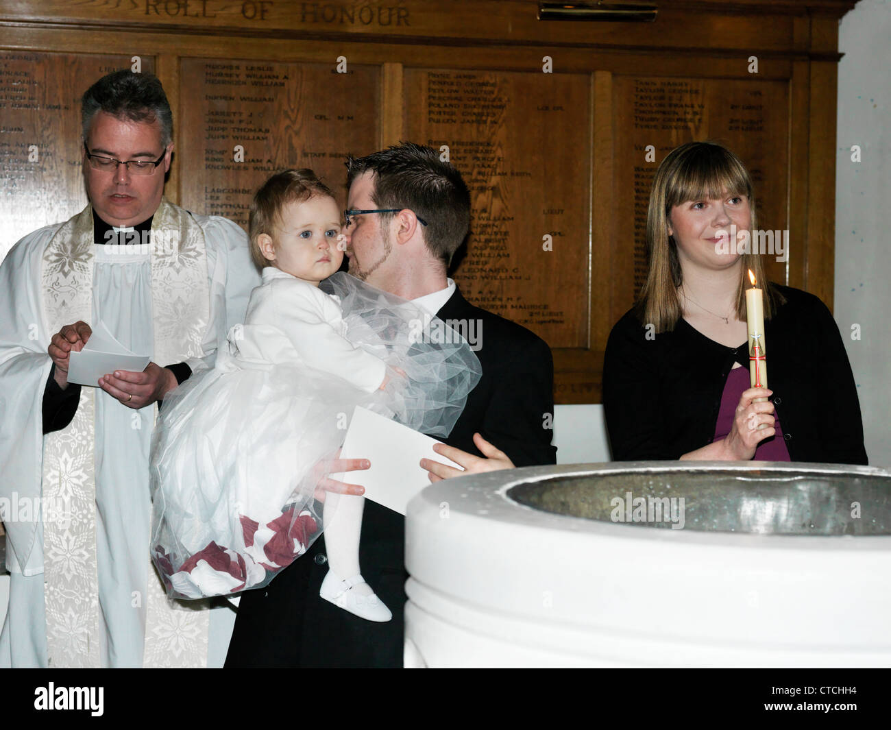Priest And Parents With Baby Girl Standing By Font With Mother Holding Candle As Presentation Of The Flame At Christening Stock Photo