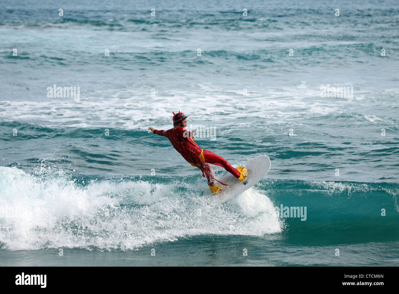Bearded man surfing a wave wearing red superhero costume. Stock Photo