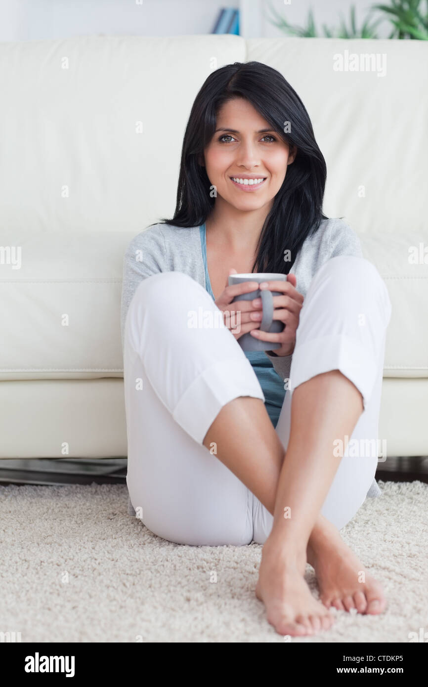 Woman holding a mug with two hands and crossing her legs while sitting on the floor Stock Photo