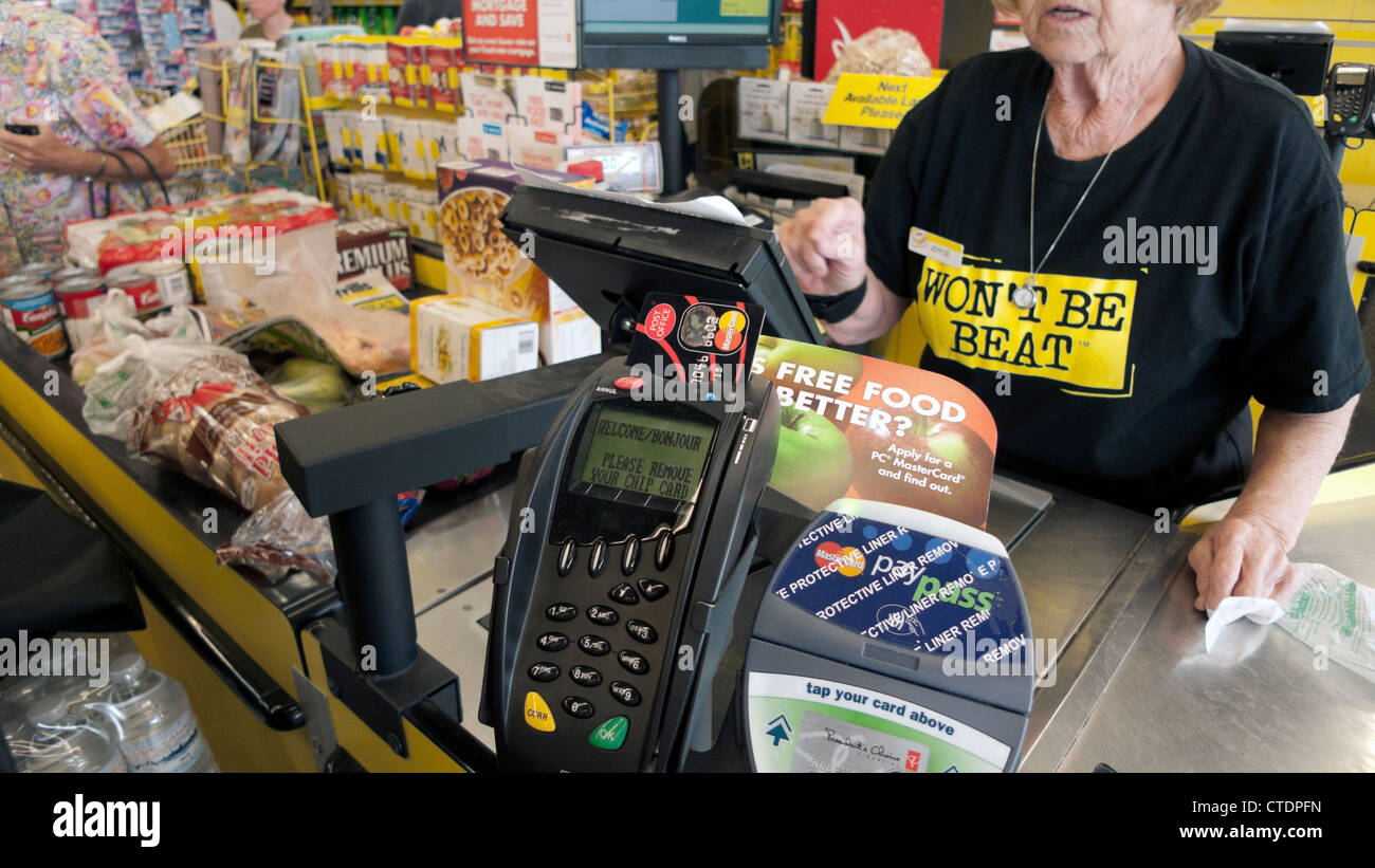 An elderly female employee older worker woman working behind the till checkout counter Jesse & Kelly's No Frills supermarket Canada   KATHY DEWITT Stock Photo