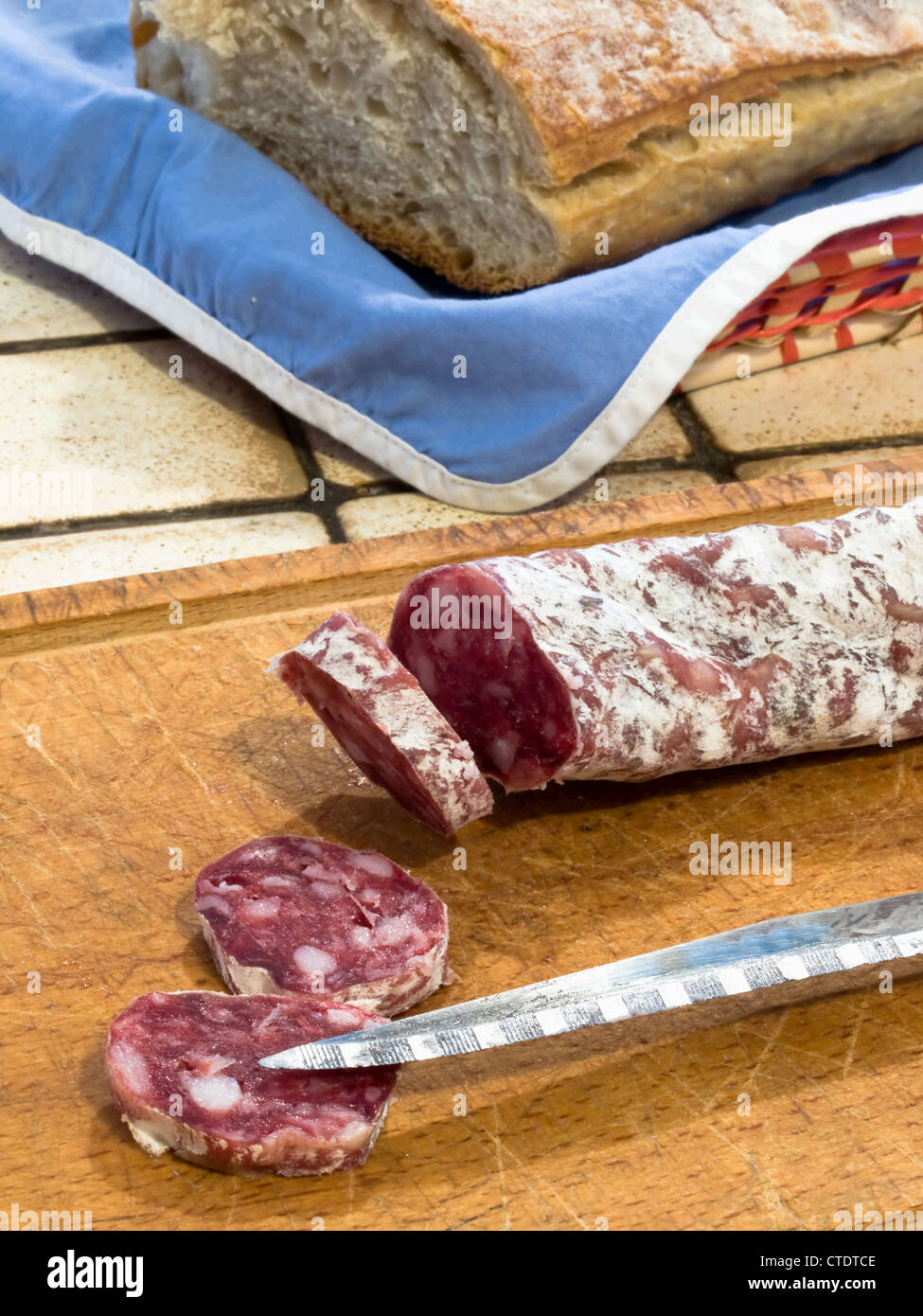 French dry sausage on a wooden cutting board with slices cut off and a knife. A bread is present in background. Stock Photo