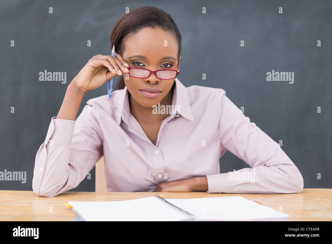 Serious teacher sitting at desk while touching her glasses Stock Photo ...