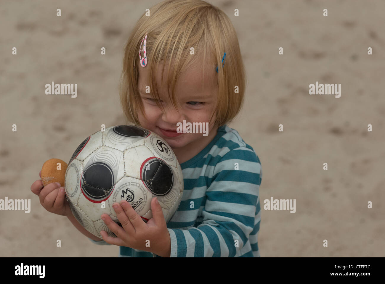 A cheerful blond baby girl is holding a toy soccer ball on sand beach