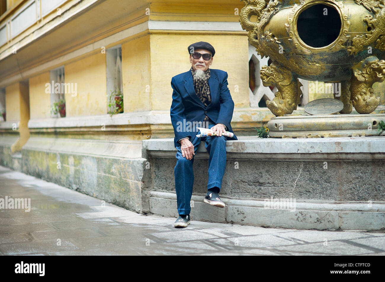 An old man in a blue suit, wearing an artist hat and sunglasses at a temple in Vietnam. Stock Photo