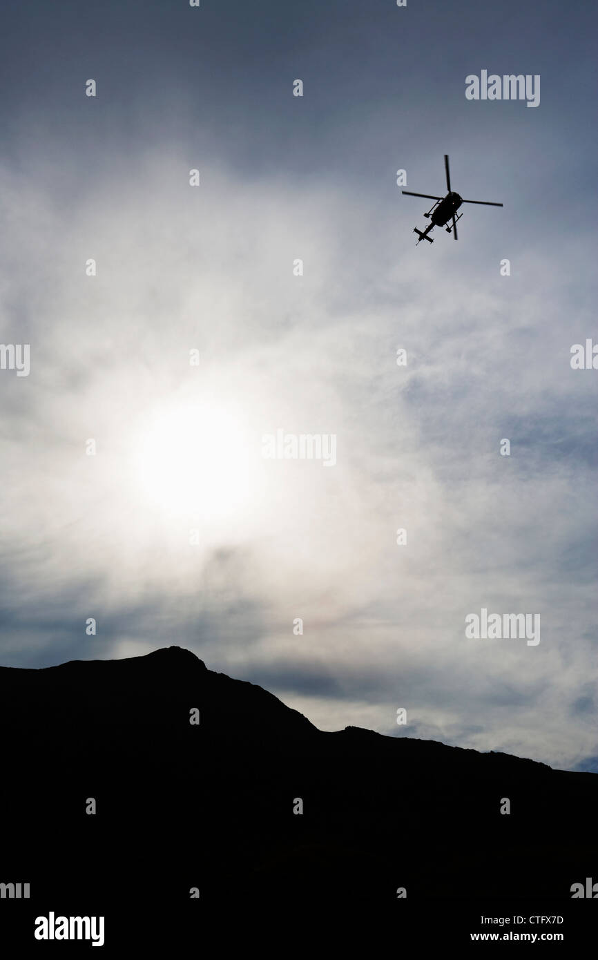 View of a mountain rescue helicopter and mountain range silhouetted against the sky and sun. Stock Photo