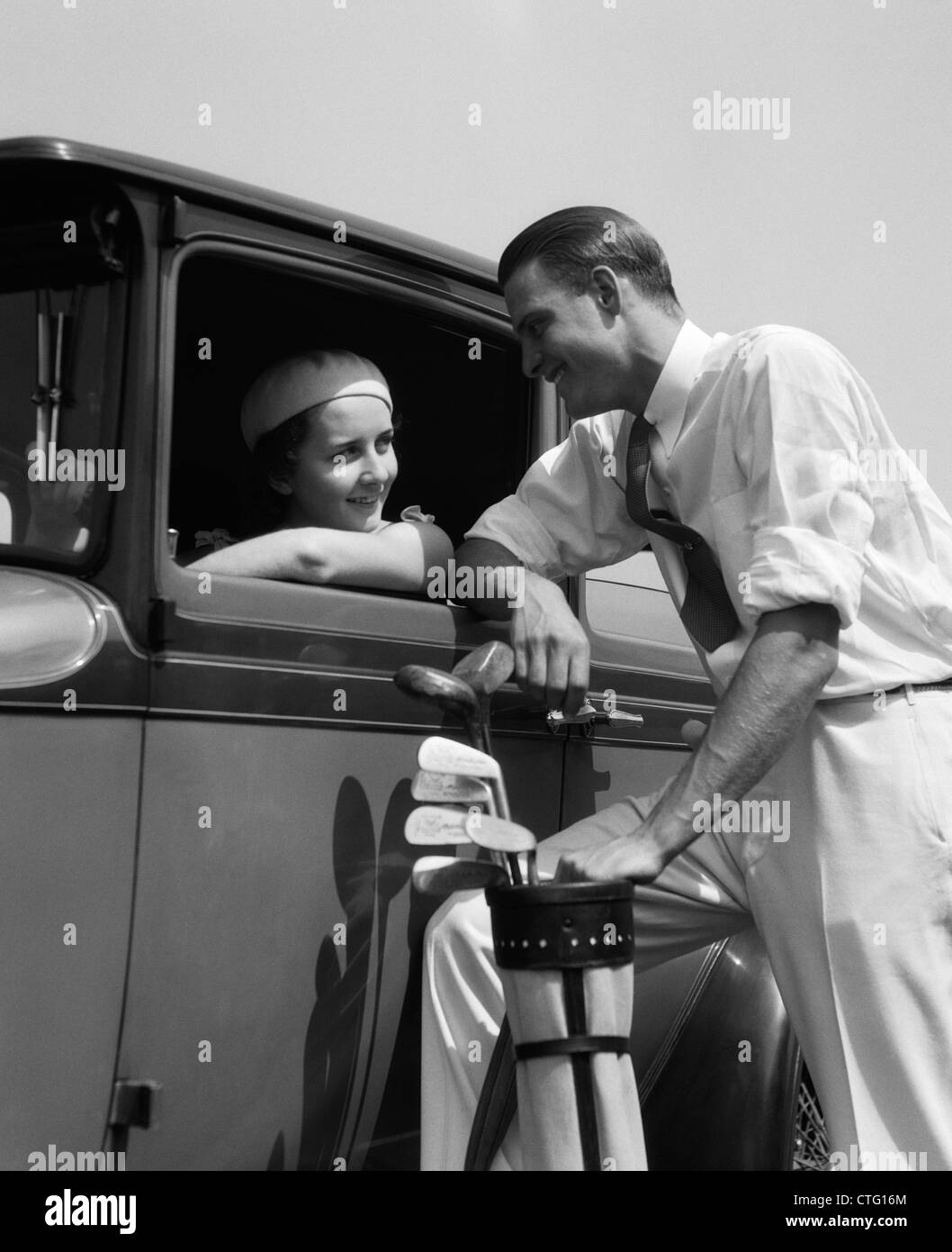 1930s WOMAN IN DRIVER'S SEAT OF CAR LEANING OUT OF WINDOW TALKING TO MAN STANDING HOLDING GOLF BAG Stock Photo