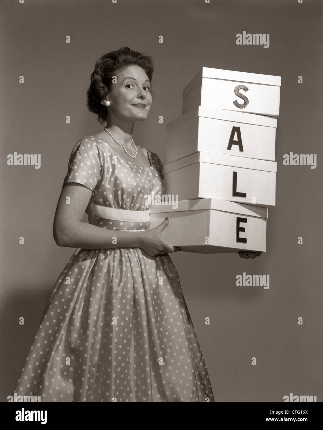 1960s WOMAN IN POLKA-DOT DRESS LOOKING AT CAMERA HOLDING A STACK OF 4 BOXES WITH ONE CHARACTER CENTERED ON EACH SPELLING SALE Stock Photo