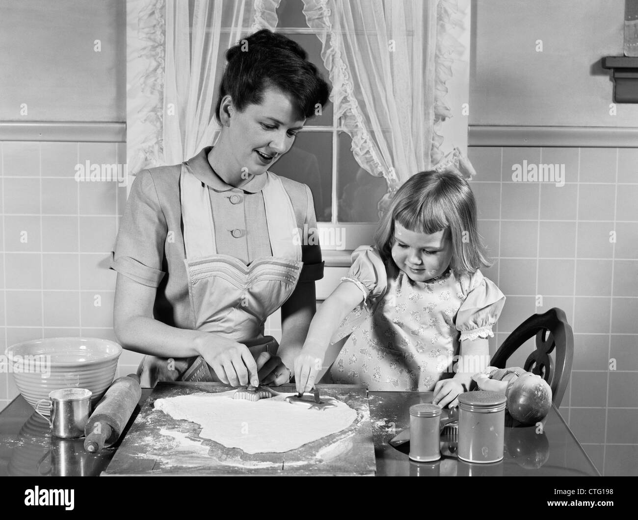 1950s MOTHER & DAUGHTER BAKING TOGETHER IN KITCHEN CUTTING DOUGH WITH COOKIE CUTTERS Stock Photo