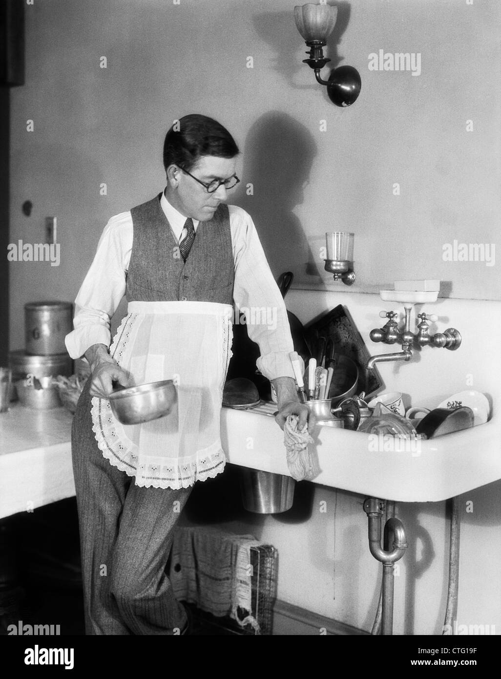 1920s MAN IN APRON LEANING ON SINK FULL OF DIRTY DISHES RAG IN ONE HAND POT IN OTHER Stock Photo