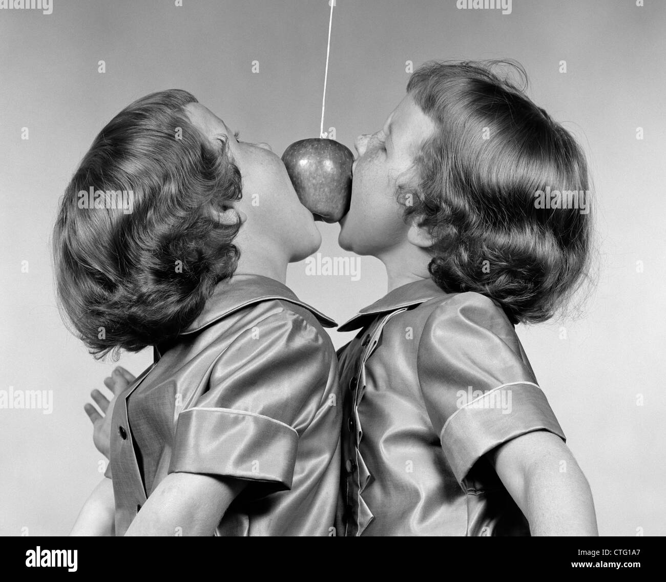 1950s IDENTICAL TWIN GIRLS TRYING TO EAT AN APPLE HANGING IN THE AIR FROM A STRING Stock Photo