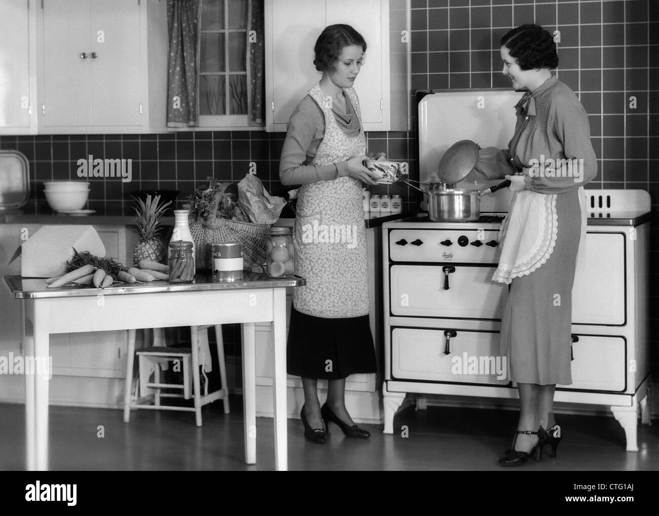 1930s WOMAN HOUSEWIFE AND FRIEND WEARING APRON COOKING FOOD IN KITCHEN ON GAS STOVE INDOOR Stock Photo