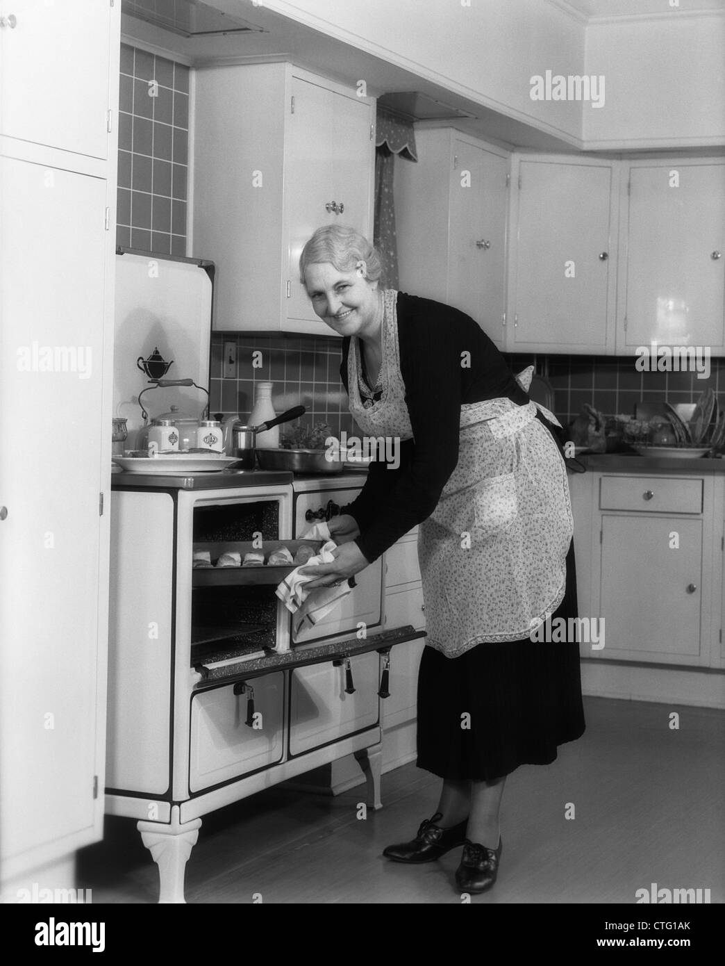 1940s ELDERLY WOMAN TAKING PAN OF ROLLS OUT OF OVEN LOOKING AT CAMERA Stock Photo