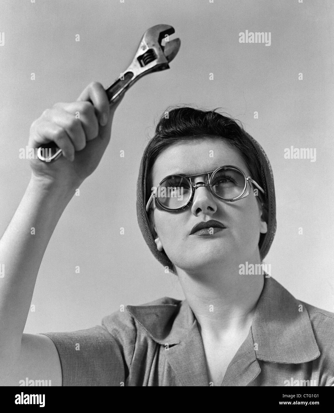 1940s ROSIE THE RIVETER WEARING SAFETY GLASSES HOLDING UP MONKEY WRENCH Stock Photo