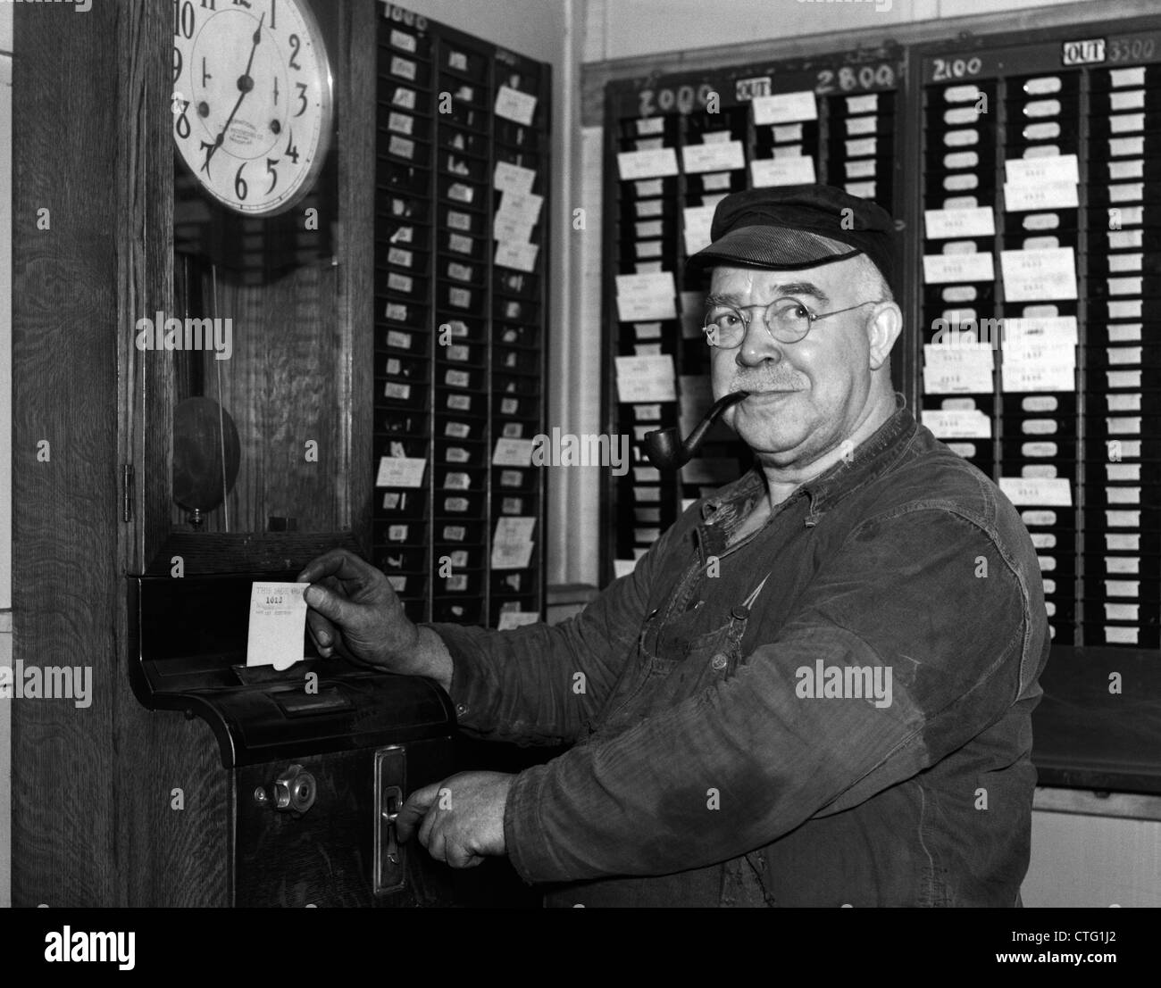 1930s ELDERLY FACTORY WORKER IN OVERALLS SMOKING PIPE PUNCHING TIME CARD Stock Photo