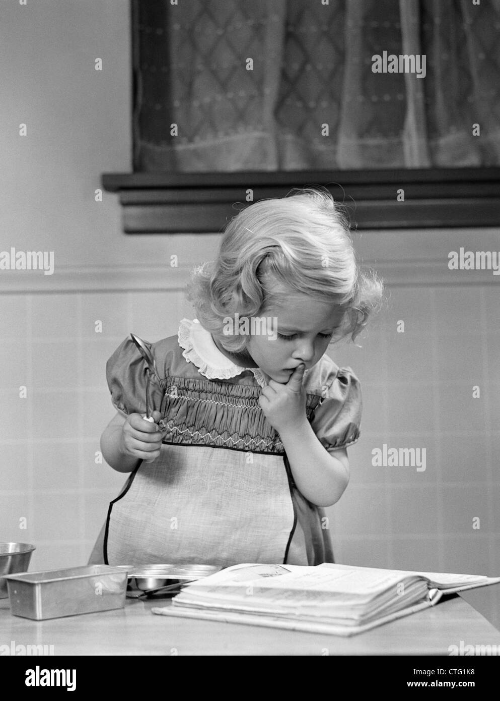 1940s LITTLE GIRL  PLAYING IN KITCHEN WEARING APRON LOOKING IN COOKBOOK GETTING READY TO BAKE Stock Photo