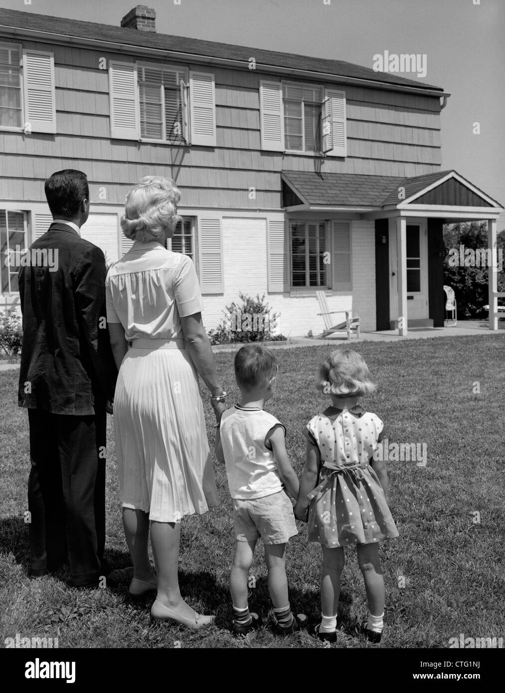 1950s 1960s FAMILY FATHER MOTHER SON DAUGHTER STANDING TOGETHER LOOKING AT SUBURBAN HOUSE Stock Photo