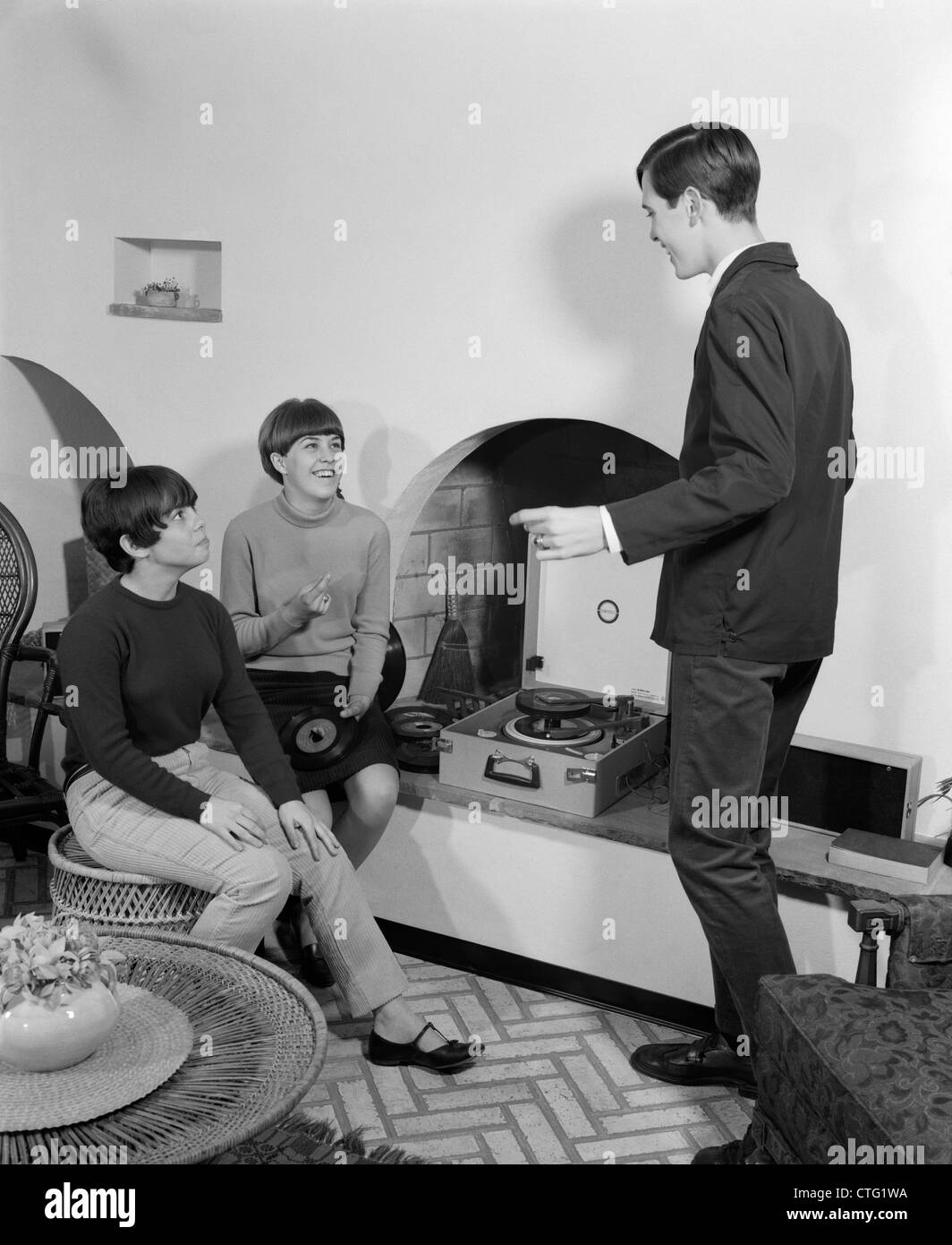 1960s 2 TEEN GIRLS SEATED ON WICKER STOOL AT FIREPLACE CHANGING 45 RPM RECORDS WITH TEEN BOY DANCING Stock Photo