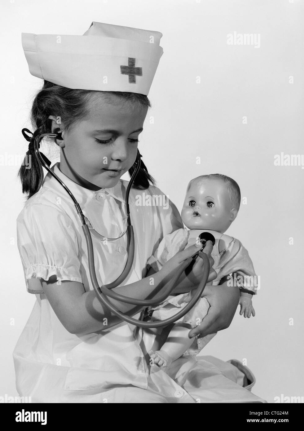 1960s GIRL IN NURSE UNIFORM HOLDING STETHOSCOPE TO BABY DOLL CHEST INDOOR Stock Photo