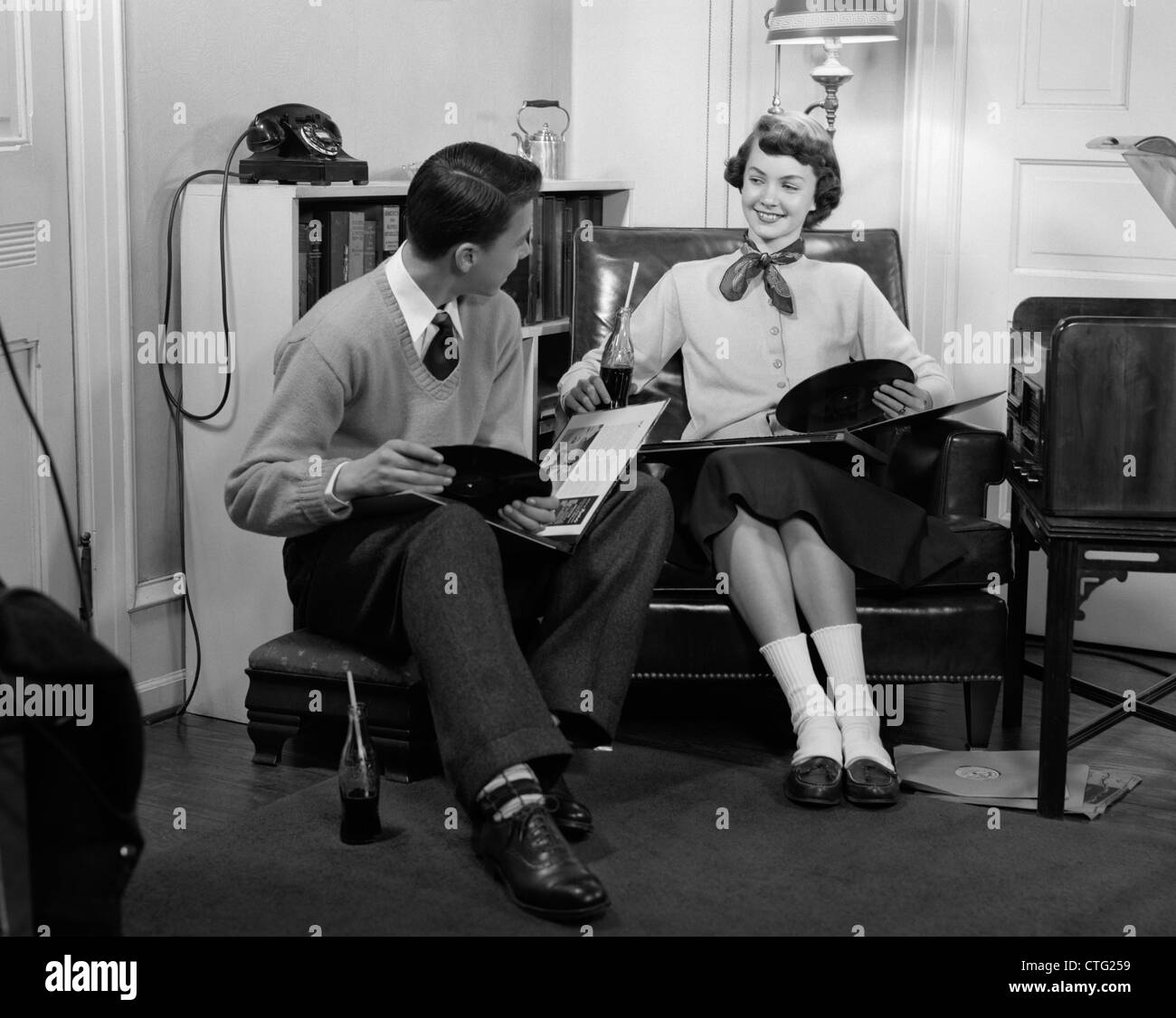 1950s TEEN BOY & GIRL SITTING IN LIVING ROOM DRINKING SODA & LISTENING TO RECORDS Stock Photo