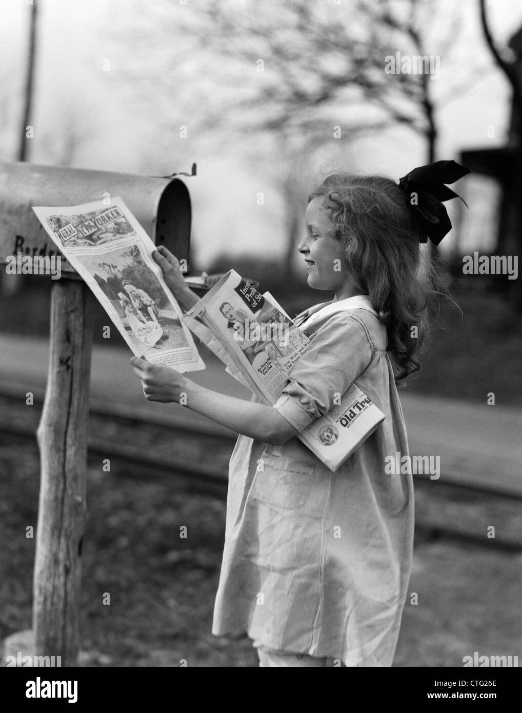 1930s 1940s LITTLE GIRL WITH BOW IN HAIR SMILING TAKING MAGAZINES OUT OF RURAL MAILBOX Stock Photo