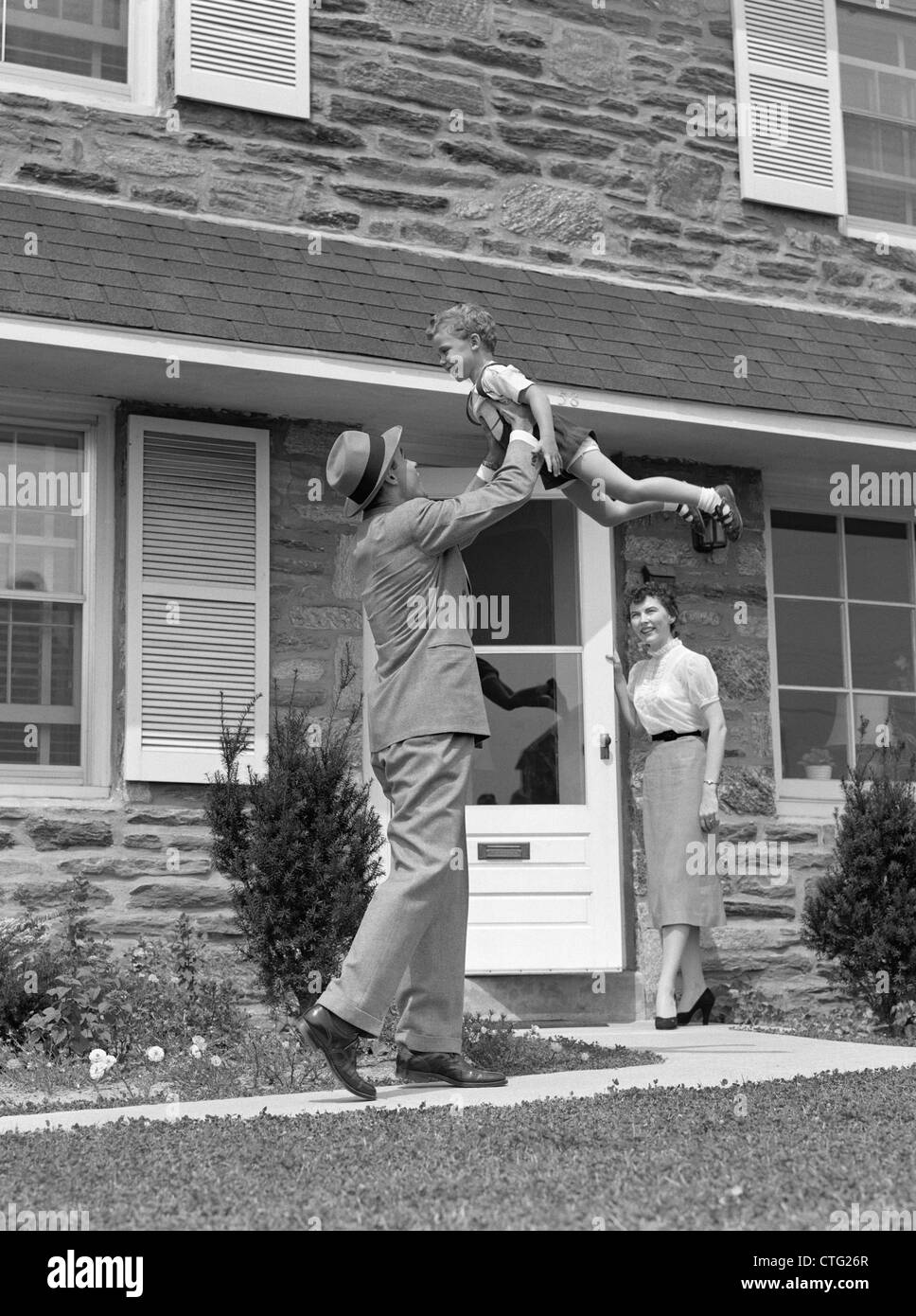 1950s 1960s FATHER COMING HOME LIFTING SON IN AIR WHILE WIFE LOOKS ON FROM DOORWAY Stock Photo