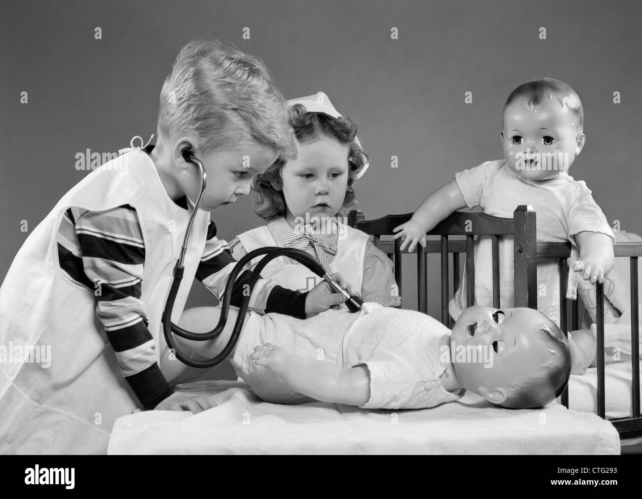 1950s BOY AND GIRL PLAYING DOCTOR AND NURSE WITH STETHOSCOPE AND DOLLS Stock Photo