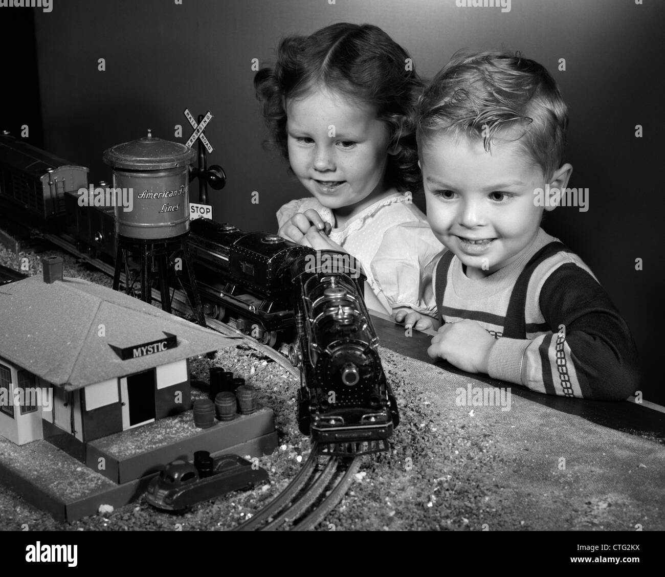 1950s BOY & GIRL STANDING AT TABLE WATCHING TOY TRAIN GOING AROUND TRACK Stock Photo