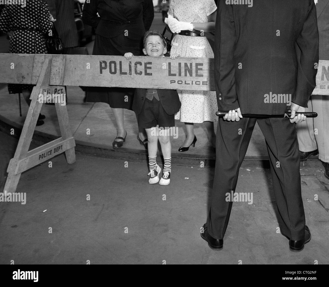 1950s BOY LOOKING OVER POLICE BARRICADE AT POLICE OFFICER WHOSE BACK IS TO THE CAMERA CROWD OF PEOPLE IN BACKGROUND OUTDOOR Stock Photo