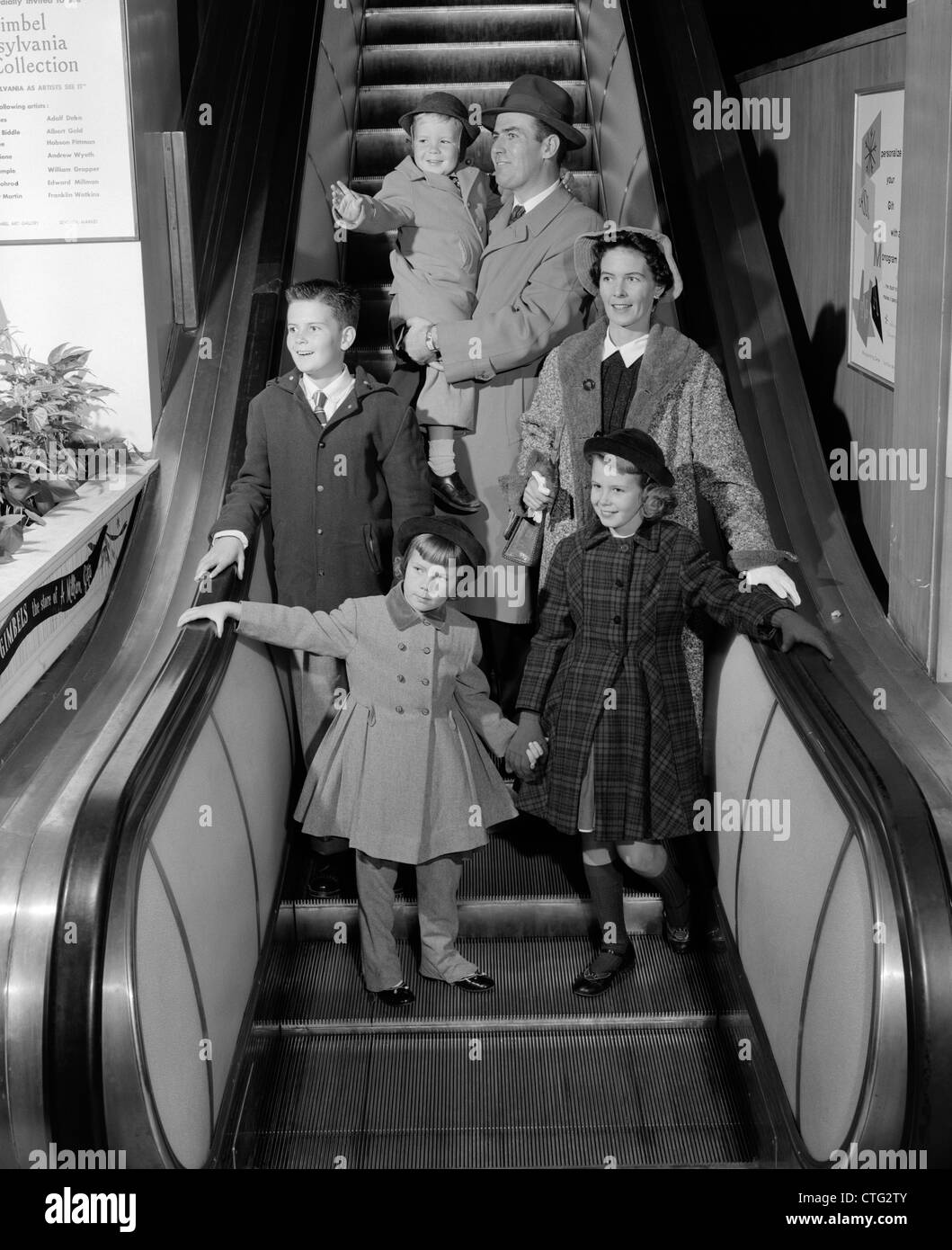 1950s SMILING FAMILY OF 6 IN WINTER COATS GOING DOWN STORE ESCALATOR Stock Photo