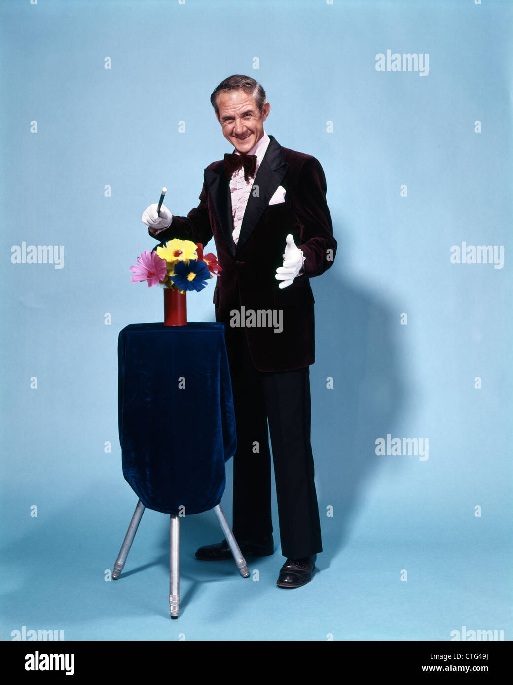 1970s FULL LENGTH SMILING MAGICIAN WAVING WAND OVER FLOWERS  LOOKING AT CAMERA Stock Photo