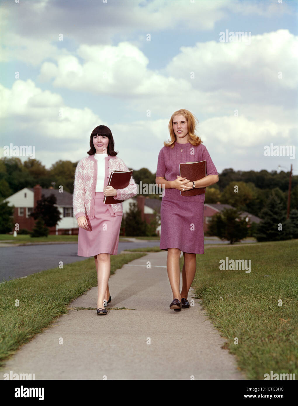 1960s TWO TEEN GIRLS CARRYING BOOKS WALKING DOWN SIDEWALK Stock Photo