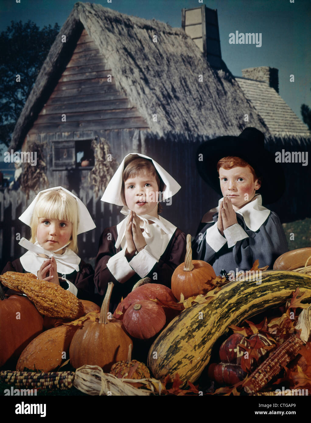 1960s THREE CHILDREN WEARING PILGRIM OUTFITS AND SAYING GRACE BEFORE THANKSGIVING HARVEST VEGETABLES Stock Photo