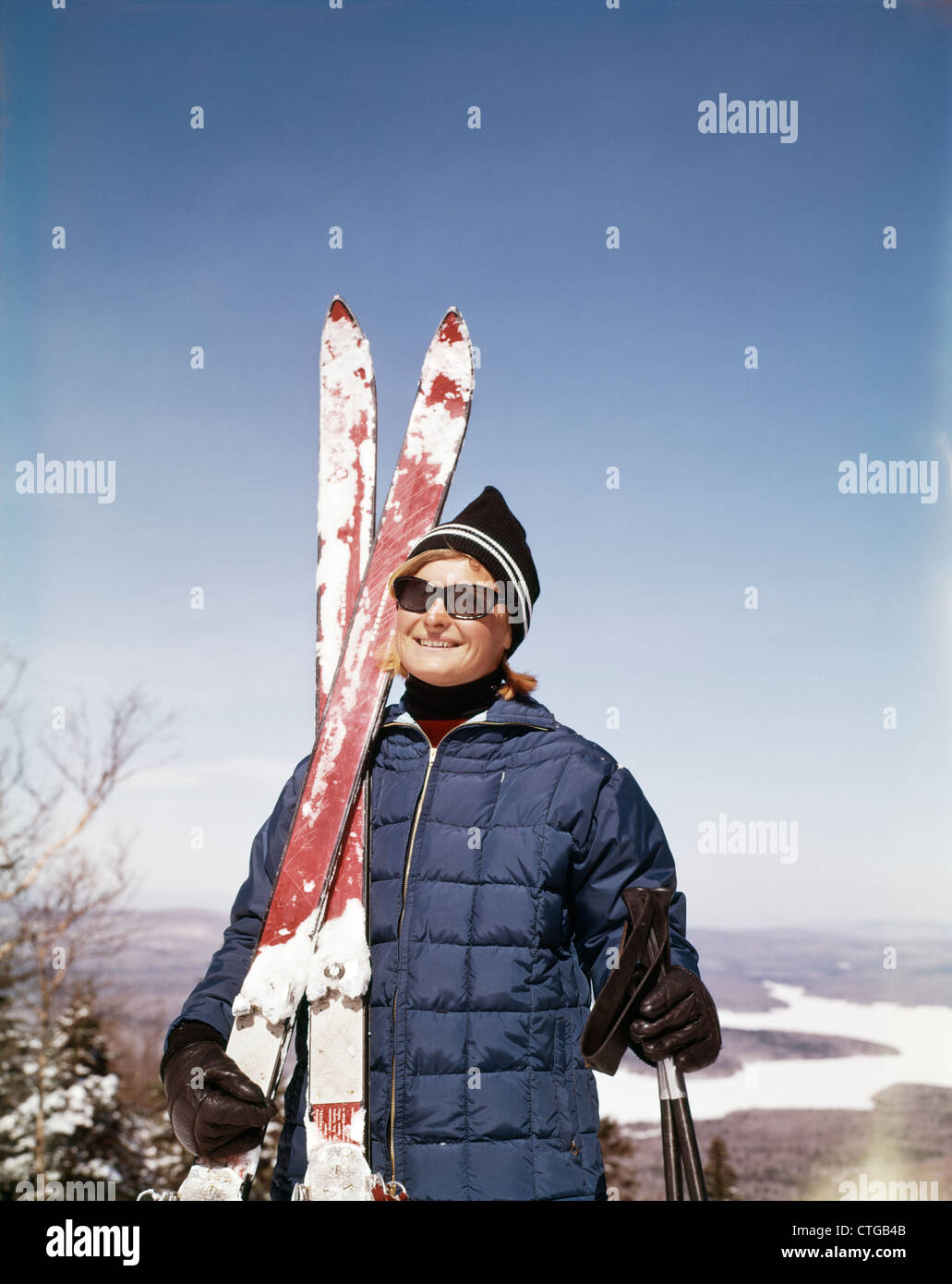 1960s PORTRAIT SMILING WOMAN OUTDOORS WEARING QUILTED DOWN JACKET SUNGLASSES HOLDING SKIS Stock Photo