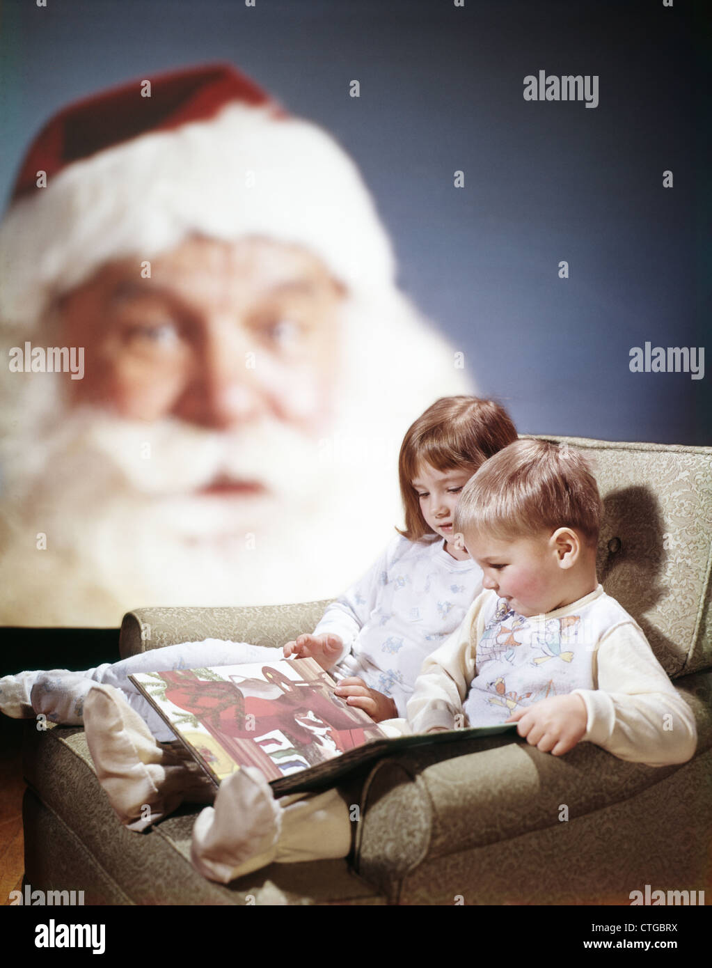 1970s CHILDREN READING CHRISTMAS STORY BOOK SANTA CLAUS IN BACKGROUND Stock Photo