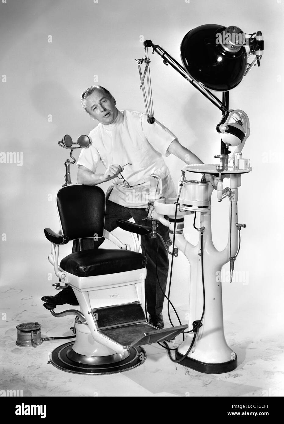 1950s MAN DENTIST HOLDING GLASSES SPEAKING FOOT PROPPED UP ON DENTAL CHAIR AND EQUIPMENT LOOKING AT CAMERA Stock Photo