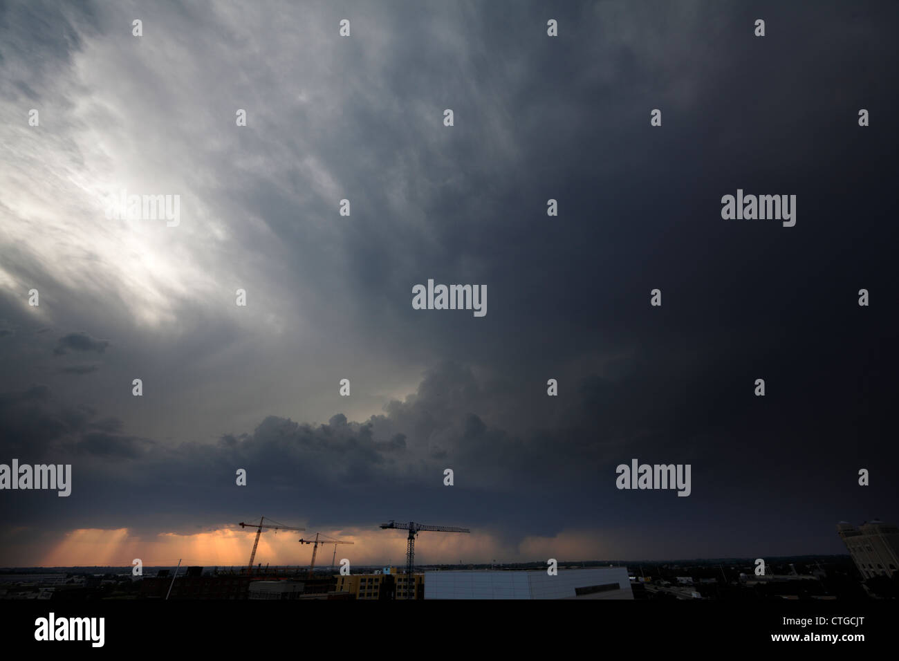 A funnel cloud lowers behind three tower cranes beyond a city. Stock Photo
