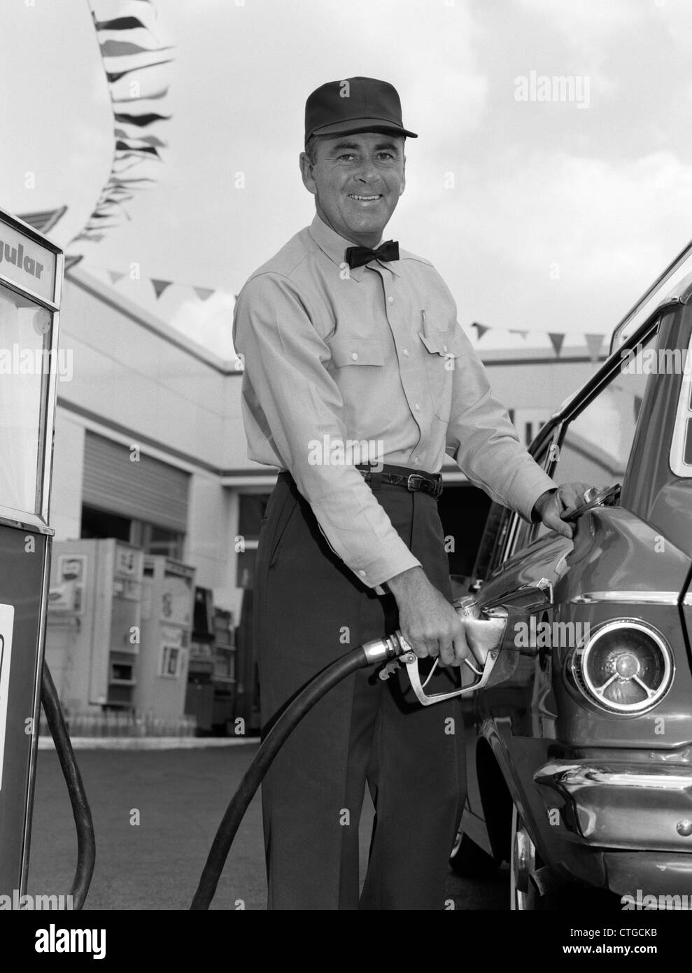 1960s MALE SERVICE STATION ATTENDANT WEARING CAP AND BOW TIE SMILING WHILE PUMPING GASOLINE OUTSIDE Stock Photo