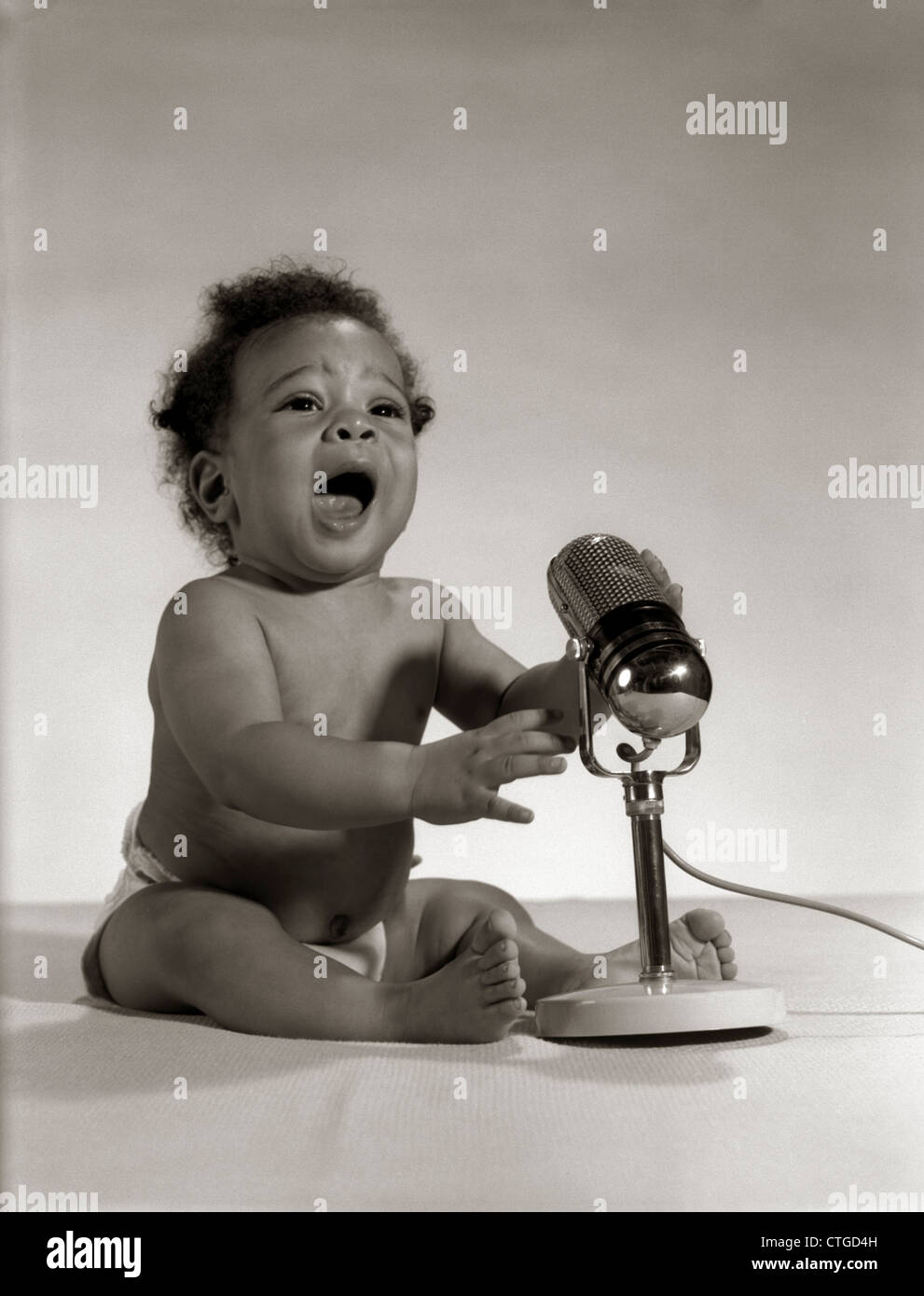 1960s AFRICAN-AMERICAN BABY SEATED IN FRONT OF OLD FASHIONED MICROPHONE WITH MOUTH OPEN BROADCASTING RADIO Stock Photo