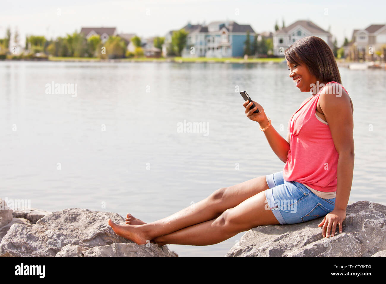 Teenage Girl Viewing Test Messages While Relaxing On The Shore Of A Residential Lake Area; Edmonton, Alberta, Canada Stock Photo