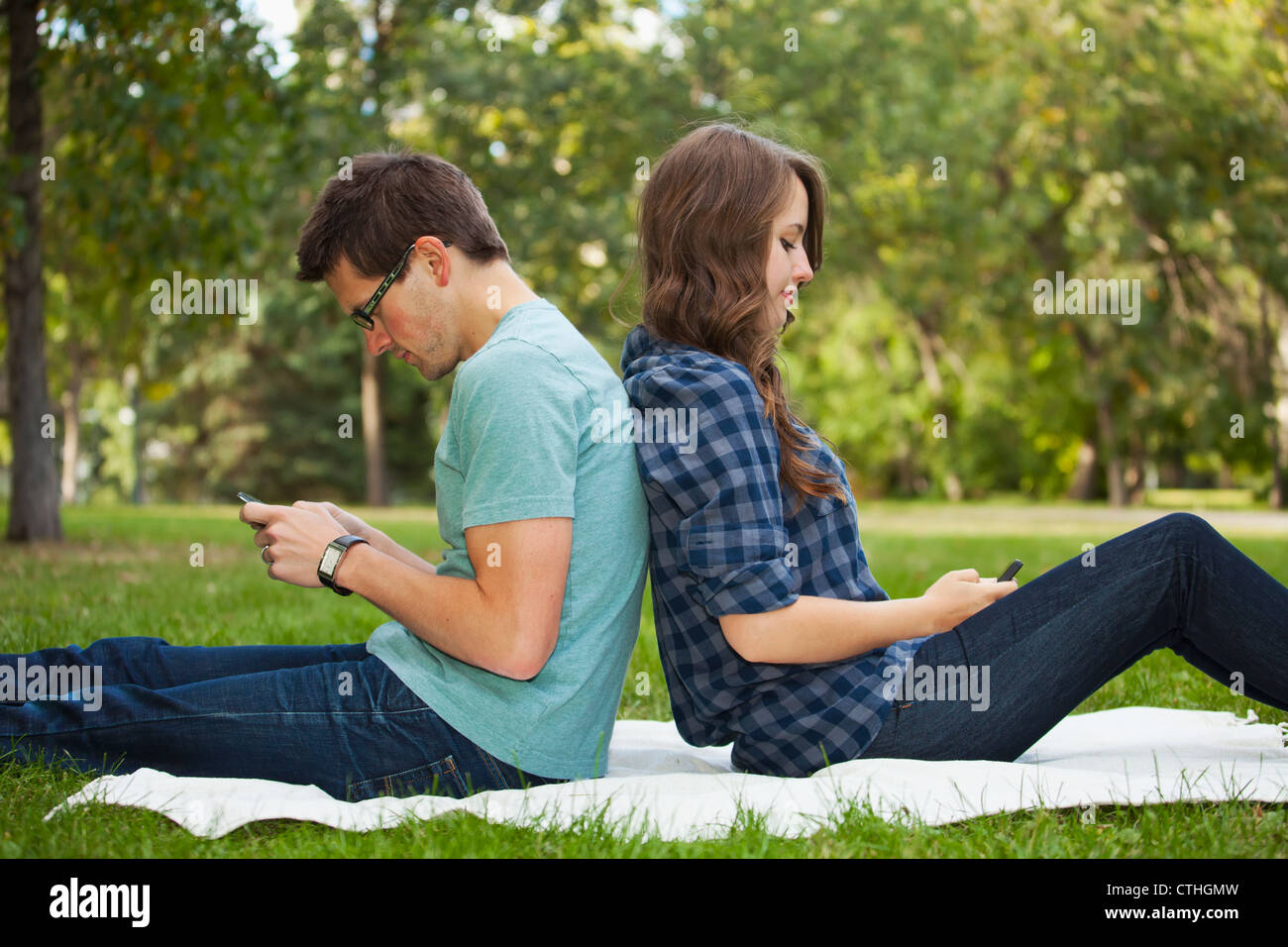 Young Couple Looking At Text Messages On Their Phones As They Sit Back To Back; Edmonton, Alberta, Canada Stock Photo